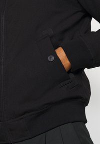 Černá bomberová bunda se zipovým uzávěrem, ribbed manžetami a boční kapsou s knoflíkovým detailem. Texturovaná tkanina, přiléhavý střih.