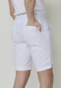 Shorts en coton blanc avec un ourlet roulé, des poches sur le devant et des poches arrière à rabat. Texture lisse avec un minimum de détails en métal.