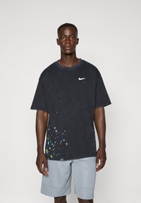 Nike oversized t-shirt i mørkeblå med blå, grønne og gule sprøjtede mønstre. Lavet af bomuldsmateriale, rund halsudskæring og korte ærmer. Sammen med lysegrå shorts.