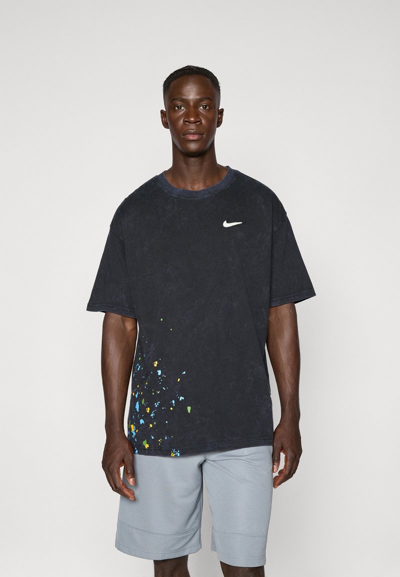 Nike oversized t-shirt i mørkeblå med blå, grønne og gule sprøjtede mønstre. Lavet af bomuldsmateriale, rund halsudskæring og korte ærmer. Sammen med lysegrå shorts.