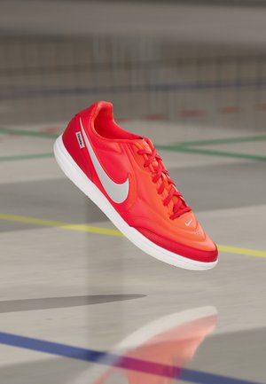 Scarpa da calcio indoor Nike Tiempo rossa e arancione con suola bianca che fluttua sopra un pavimento di palestra con linee da campo sportivo.