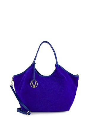 Grote blauwe suède handtas met twee handvatten, afneembare schouderriem en een ronde gouden logohanger die aan één handvat hangt.