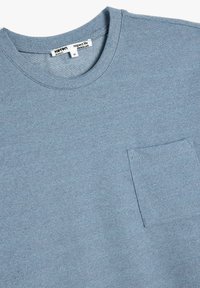 T-shirt blu con texture, collo rotondo, maniche corte e una piccola tasca sul lato sinistro del petto. Etichetta visibile nell'area del colletto.