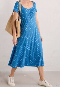 Blå klänning med prickar, med en figurnära överdel, utsvängd kjol och korta ärmar. Bärs med en vävd beige väska och vita sneakers. Slät tygyta.