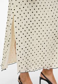 Midi kjole med polkadotter, laget av lett, teksturert stoff i kremfarge med svarte prikker, sidestilt splitt og bølget nederkant. Sorte høye hæler er synlige.