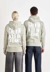 Due individui indossano felpe verde chiaro con ampio testo bianco: "CALVIN KLEIN JEANS." Le felpe hanno tasche a marsupio e polsini a costine.
