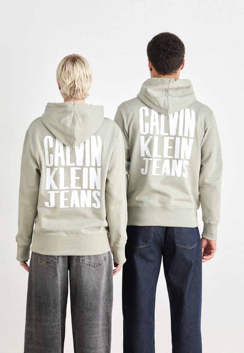 Due individui indossano felpe verde chiaro con ampio testo bianco: "CALVIN KLEIN JEANS." Le felpe hanno tasche a marsupio e polsini a costine.