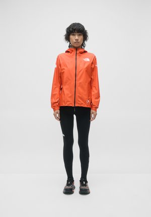 Veste imperméable orange vif avec fermeture éclair intégrale, manches longues et accents de logo, associée à des leggings noirs et des chaussures techniques.