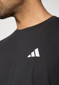 Juodas trumpomis rankovėmis marškinėlis, pagamintas iš minkštos medžiagos, su balta logotipo dizainu, turinčiu tris kylancias juostas kairėje krūtinės pusėje.