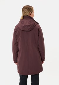 Vrouw met blond haar in een lage paardenstaart, gekleed in een lange, met een capuchon maroon winterjas en zwarte broek, met haar rug naar de camera gekeerd tegen een witte achtergrond.