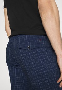 Tmavě modré kostkované kalhoty s kontrastními světle modrými pruhy, se zadní kapsou na knoflíkový uzávěr a malým logem.