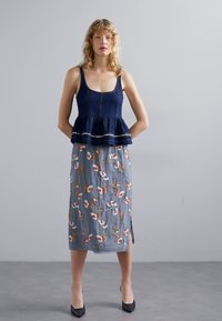 Marineblaues Peplum-Top mit dünnen Trägern, kombiniert mit einem grauen, bestickten Midirock mit floralen Mustern und Seitenschlitzen. Schwarze High Heels.