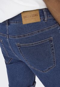 Jeans in denim di colore blu scuro con una texture liscia, dotati di un'etichetta in pelle in vita e doppie tasche posteriori con cuciture a contrasto.