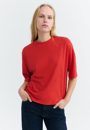 Blonde Frau mit langen Haaren, die ein lockeres rotes kurzärmeliges Oberteil und dunkelblaue Jeans trägt, steht mit den Händen in den Taschen.