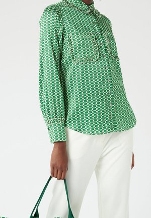 Person wearing a green patterned button-up shirt with pockets, white pants, holding a green-handled bag against a plain background.
