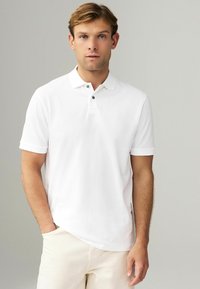 Hvid polo shirt med korte ærmer, to-knaps placket og broderet logo på brystet. Modellen bærer lyse bukser.