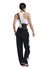 Svart axelremsväska med en rem, bärs över en vit linnen, ihop med svarta byxor med vita sidostreck, sedd bakifrån.