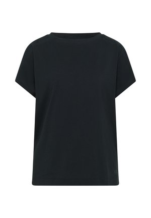Mujer sonriendo, lleva una camiseta oscura de manga corta, shorts beige, zapatos sin cordones color tostado, sostiene un bolso grande de mimbre y está de pie en interiores.