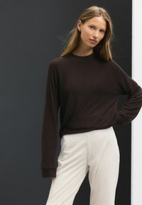 Suéter marrón oscuro con cuello redondo, mangas largas y puños acanalados, combinado con pantalones de textura suave y color claro. Diseño minimalista.