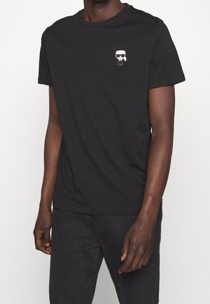 T-shirt en coton noir avec un col rond, des manches courtes et un petit emblème de personnage de dessin animé blanc sur la poitrine gauche.