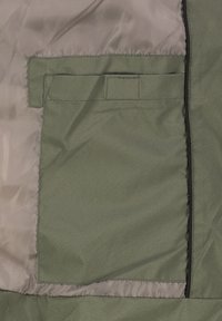 Binnenkant van een olijfgroene jas met een rechthoekige zak met klep, een gladde voering en een contrasterende zwarte ritssluiting langs de rand.