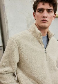 Pull en polaire beige avec un design à demi-zip et un col haut. Tissu doux et texturé, associé à une chemise grise en dessous.