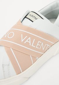 Baskets blanches avec un élastique rose clair portant le texte "VALENTINO". Tige en cuir lisse, bout arrondi et semelle en caoutchouc texturée.
