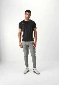 Camiseta atlética preta com detalhes dourados, combinada com calças de treino cinzentas e sapatilhas brancas. Design ajustado com mangas curtas e painéis laterais.