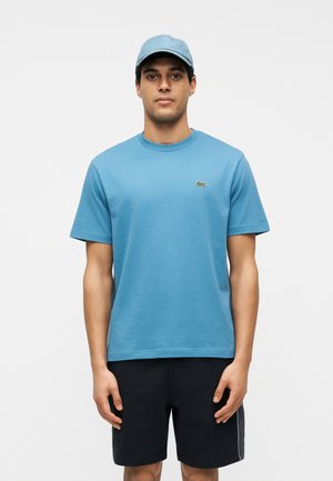 Άνδρας που φοράει ανοιχτό μπλε καπέλο, γαλάζιο κοντομάνικο μπλουζάκι με μικρό λογότυπο και μαύρο σορτς, στέκεται μπροστά σε καθαρό λευκό φόντο.