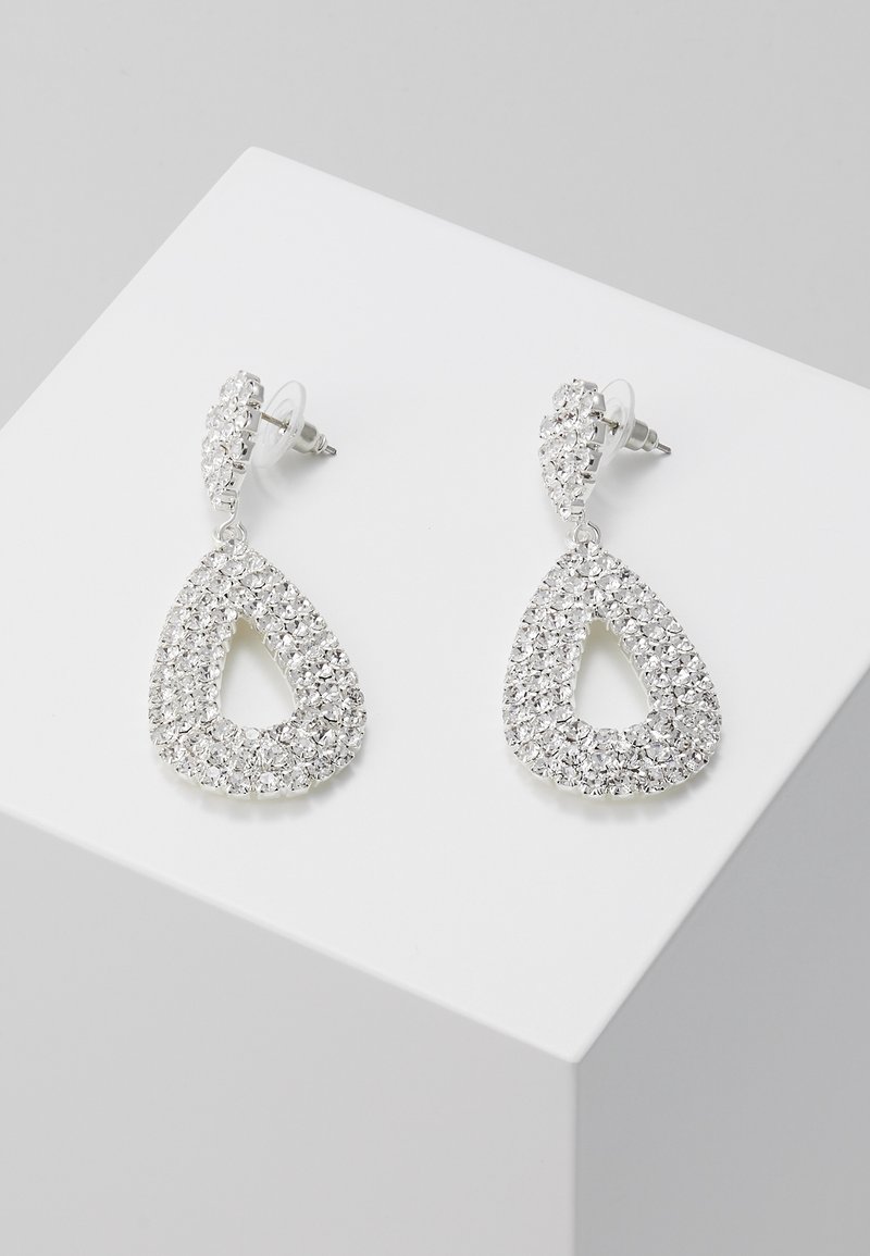 Boucles d’oreilles pendantes couleur argent avec un design ouvert en forme de goutte, incrustées de strass clairs, présentées sur une surface blanche inclinée.
