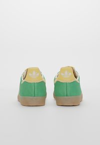 Gröna mockaskor med en gul hälflik, beige detaljer och brun gummisula. Har dekorerande sömmar längs hälen.