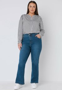 Jeans de mezclilla acampanados en azul medio con una cintura alta, combinados con una blusa de rayas en blanco y negro con cuello mao. Zapatillas blancas.