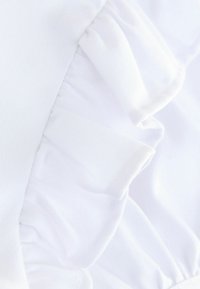 Vitt tyg med volangdetaljer längs kanten, med en slät, texturerad yta och subtil lyster.