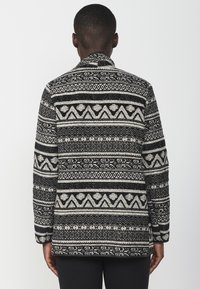 Schwarz-weiß gemusterte Jacke mit Kragen, die geometrische Designs und strukturierten Stoff aufweist. Lange Ärmel und gerader Schnitt.