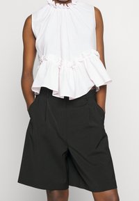 Top sin mangas texturizado blanco con un cuello fruncido y un dobladillo con volantes, combinado con pantalones cortos negros ajustados que tienen bolsillos laterales.