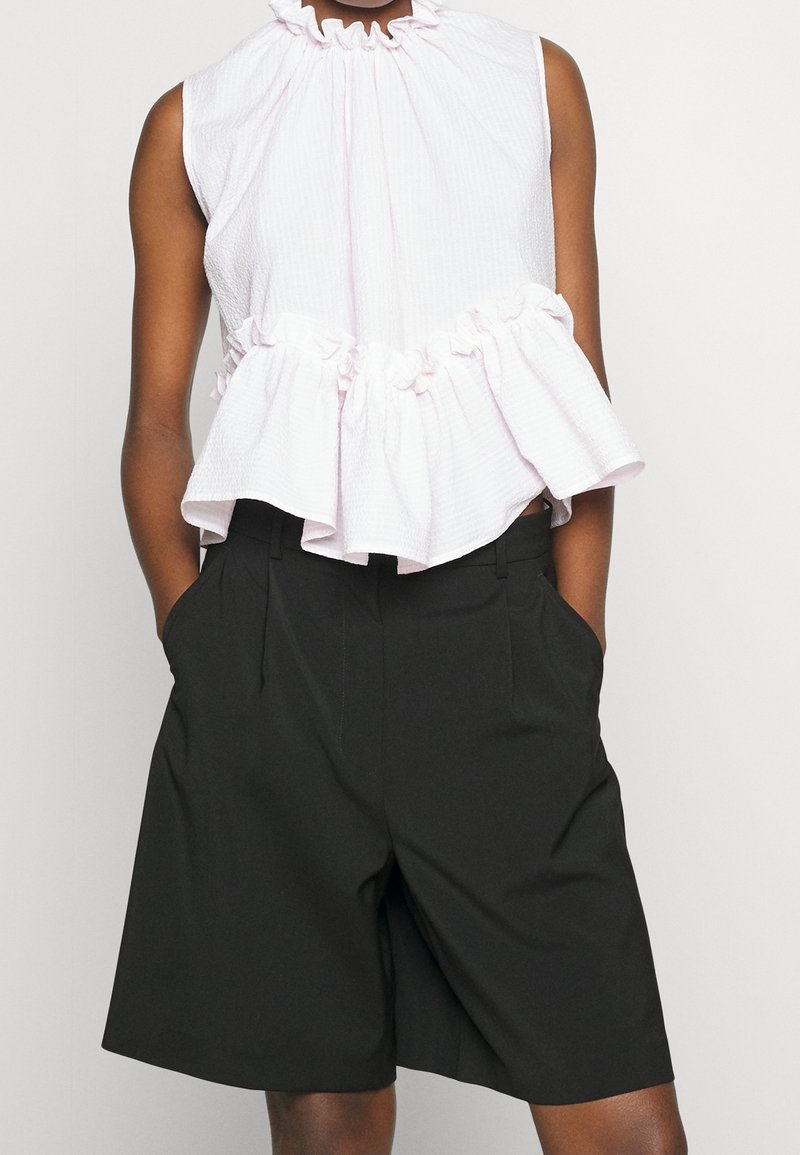 Top sin mangas texturizado blanco con un cuello fruncido y un dobladillo con volantes, combinado con pantalones cortos negros ajustados que tienen bolsillos laterales.