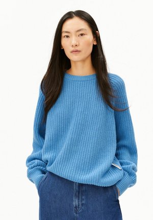 Frau mit langen dunklen Haaren, die einen blauen gestrickten Pullover und blaue Jeans trägt, steht mit den Händen in den Taschen vor einem weißen Hintergrund.