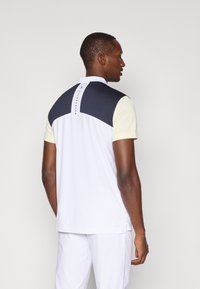 Koszulka polo z krótkim rękawem w kolorze białym z granatowym i kremowym wzorem w blokach kolorystycznych. Posiada kołnierzyk i guziki. Materiał wydaje się lekki.