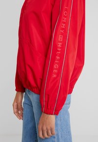 Giubbotto antivento rosso leggero con polsini ed orlo elastici, caratterizzato da una striscia bianca lungo la manica e il logo "TOMMY HILFIGER" in rilievo.