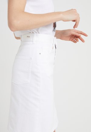 Person iført et hvidt denimskørt med en baglomme og et logo på linningen, kombineret med en hvid t-shirt på en ensfarvet baggrund.