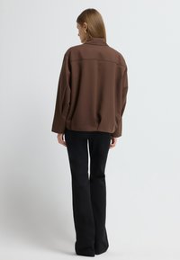 Brun, oversized knapstående jakke med krave, lange ærmer og en glat stofstruktur. Båret med sorte udvidede bukser.