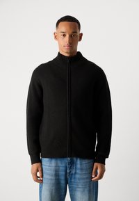 Pull noir en maille zippé avec col montant et tissu texturé, doté de longues manches et d'une coupe décontractée. Associé à un jean bleu.