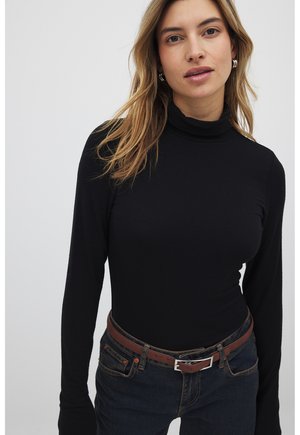 Schwarzes langärmliges Rollkragenoberteil aus weichem Stoff, kombiniert mit dunkler Jeans und einem braunen Gürtel mit silberfarbenen Beschlägen.