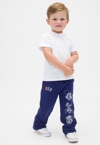 Ung dreng står iført hvid t-shirt, marineblå bukser med Disney-figurers ansigter og hvide sneakers på ensfarvet baggrund.