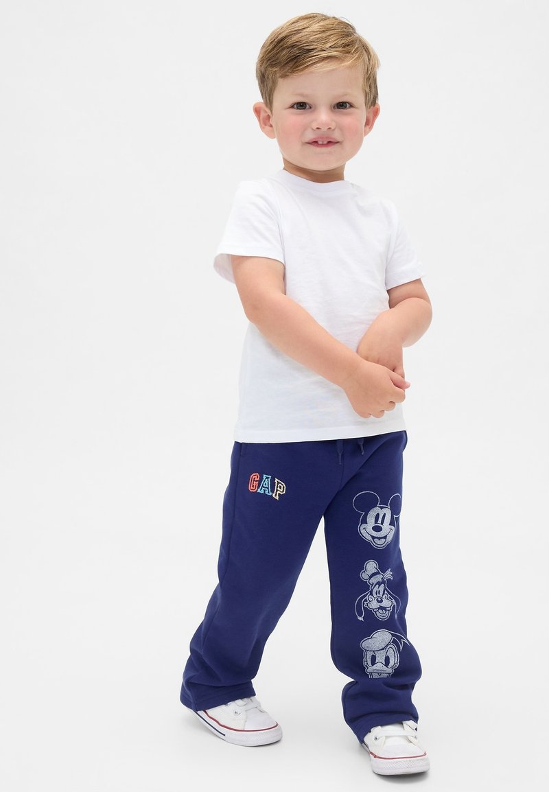 Ung dreng står iført hvid t-shirt, marineblå bukser med Disney-figurers ansigter og hvide sneakers på ensfarvet baggrund.