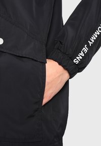 Czarna kurtka przeciwdeszczowa z zamkiem błyskawicznym, elastycznymi mankietami i boczną kieszenią. Akcentowana białym napisem na rękawie i gładką fakturą.