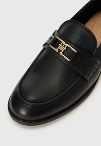Black leather loafer showcasing a smooth texture, rounded toe, and a gold-toned metal accent strap with a logo. Flat sole design.