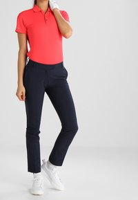 Röd polo med korta ärmar, som kombineras med marinblå byxor. Bärs med vita sneakers. Handske synlig på en hand. Enkel, sportig design.