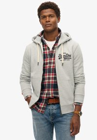Sudadera gris con cierre, con un bolsillo tipo canguro, superpuesta a una camisa de cuadros roja y azul. Presenta cordones blancos y un logo en el pecho.
