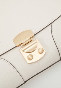 Valkoinen, teksturoitu nahkalaukku, jossa kultainen lukko. Siinä on sileä viimeistely, kohokuvioitu kuvio ja kontrastivärinen tumma nahkareunus.